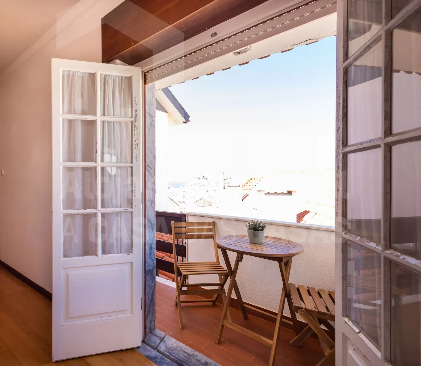 Portas de madeira abertas para a varanda, com mesa e cadeiras e vista sobre a vila