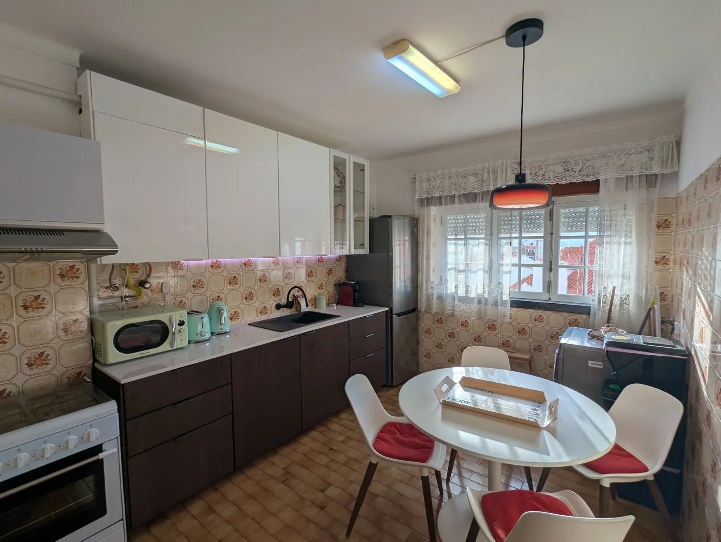 Cozinha com armários brancos, fogão, frigorífico e mesa de refeições junto à janela