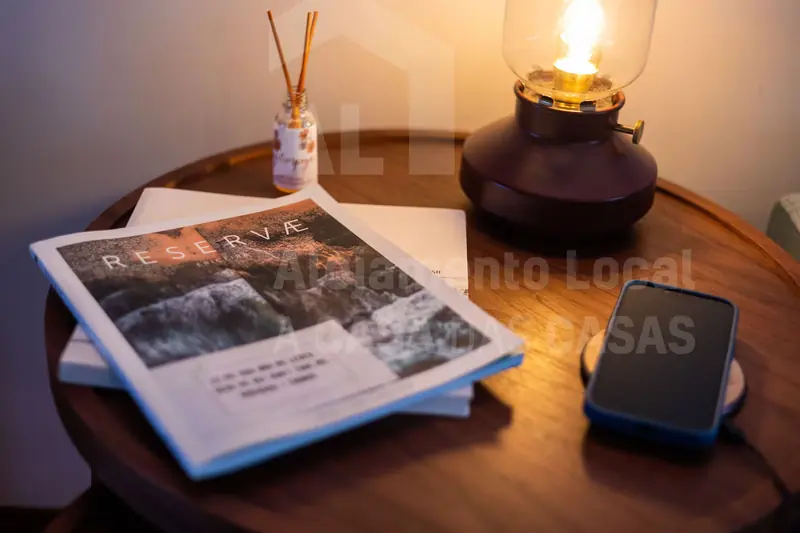Candeeiro aceso sobre uma mesa redonda de madeira, com revista e difusor de aroma